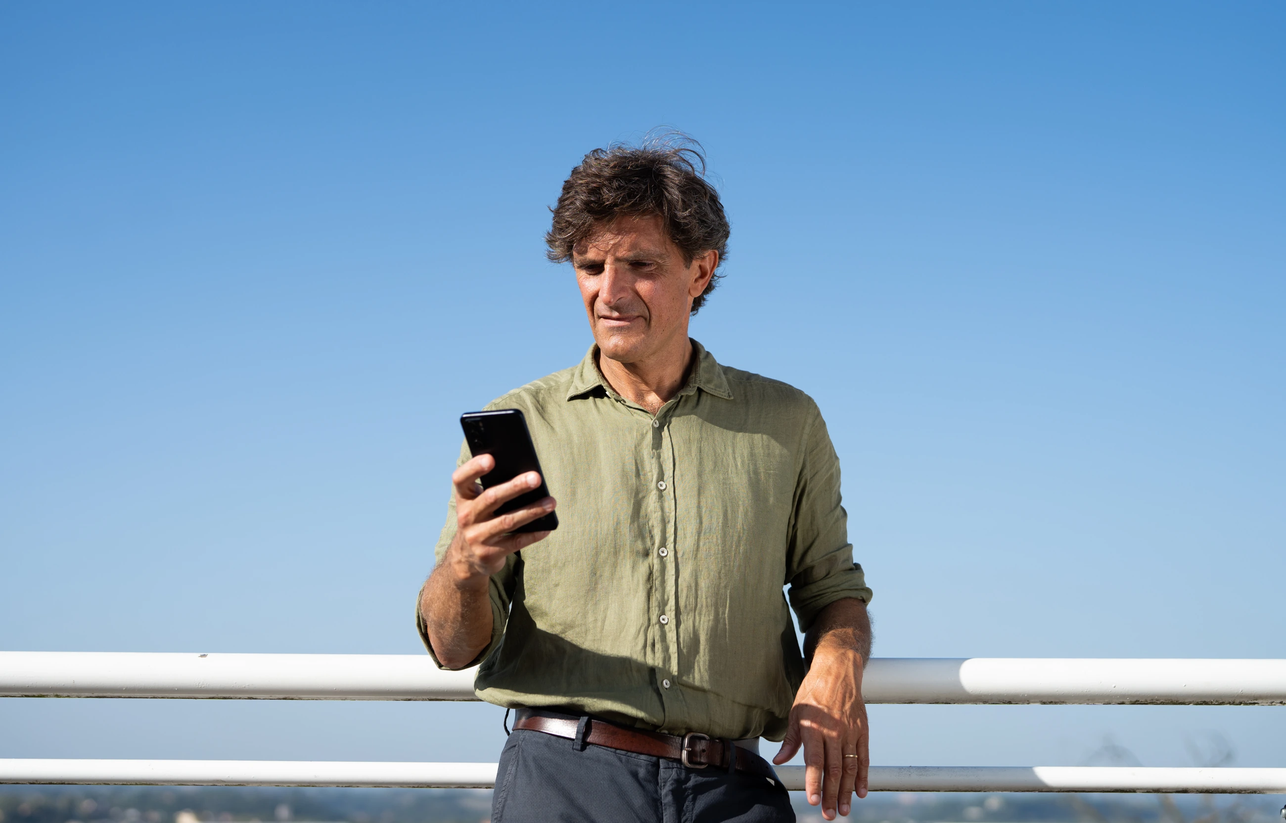 UNFCU Elite Card lifestyle portrait: a man in an olive shirt using his phone against a clear Roman sky